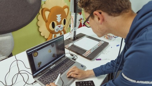 A teenage boy leans down to type instructions into a laptop.
