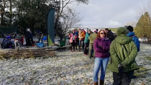 Pobl wedi’u lapio yn erbyn yr oerfel yn sefyll yn siarad mewn cae gaeafol; rhai eraill yn tynnu nwyddau allan mewn ardal wedi’i hamgáu gan ffens wiail.