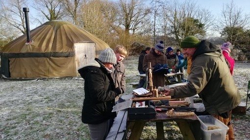 Pobl mewn cae gaeafol yn archwilio cyfarpar cerfio pren wedi’i osod ar fwrdd picnic; mae iwrt i’w weld yn y cefndir.