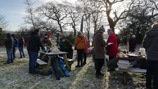 Tua phymtheg o bobl mewn siacedi gaeaf trwchus a hetiau’n sefyll o gwmpas byrddau picnic mewn cae dan haen o rew.