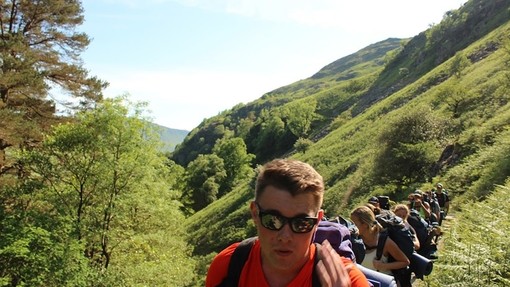 A line of young people walk along a path through a green valley on a summer day.