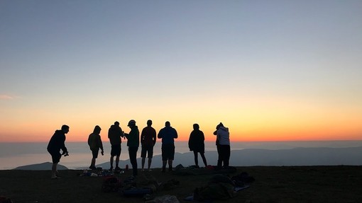 Black silhouettes of the summer school attendees standing on the edge of a mountain as the sun sets behind them.