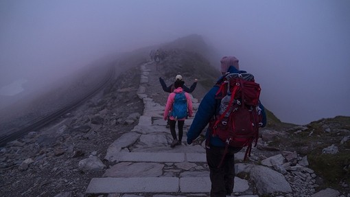 Two people wearing high-viz jackets walk along a narrow ridge into the fog.