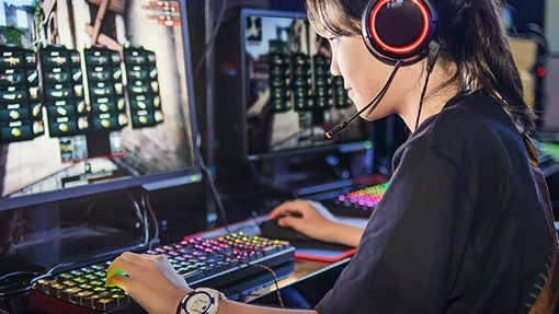 A young female student wearing headphones works at a glowing keyboard and monitor.