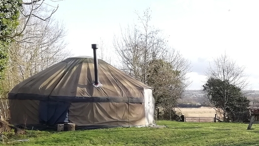A large canvas yurt in a small field; the yurt has a metal chimney; there is a seating area outside with logs and picnic benches; river meadows and the Towy itself are visible in the background. 