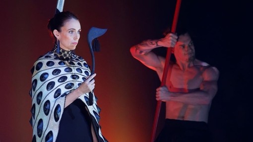 Jacinda Ardern mewn gwisg Aotearoa yn cynnwys dilledyn uchaf tebyg i bonsio â phrint, crib gwallt tebyg i lafn, a bwyell gam las addurnol.