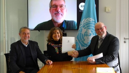 Gabriela Ramos a Steven Hartman yn siglo dwylo wrth fwrdd cynhadledd pren; mae Gustavo Merino yn eistedd wrth eu hochr tra mae Luiz Oosterbeek yn gwylio o sgrin fawr uwch eu pennau.