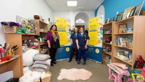 Michelle Treasure, Janet Ellis a Joanne Begley yn sefyll yn y cwpwrdd cymunedol, ystafell fach â silffoedd ar bob ochr yn dal teganau, llyfrau, offer cegin, a nwyddau eraill i’r cartref.