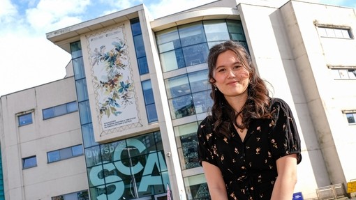 A smiling student standing in front of a giant canvas on the top of a University building in Swansea which features her work.