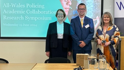 Three smiling people standing in front of a desk with a large screen behind them.