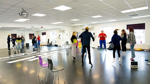 A circle of seven students jump in synch in a dance rehearsal studio with mirrored wall.