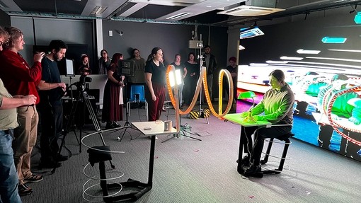 In the immersive room, a student sits at a small desk; green light from a desk lamp shines across her hands and face; thirteen people stand across from her watching, while one man films using a tripod camera.