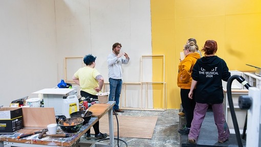 Four students listen to a tutor while standing in the middle of a set building workshop.