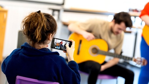 Using her phone, a young woman films a male student playing acoustic guitar.