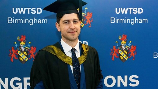 Ivan Naydenov in graduation gown in front of blue stand with UWTSD logo