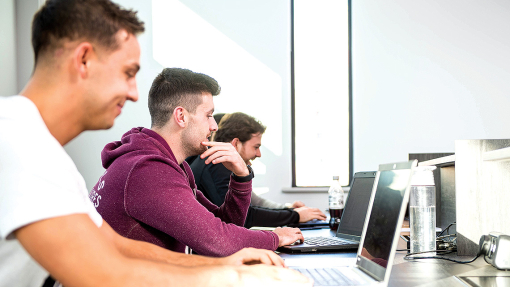 A row of students work on their laptops on a long desk in the Fforwm Library.