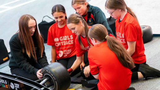 A group of girls dressed in red hoodies look closely at the wheel of a motorsports vehicle.