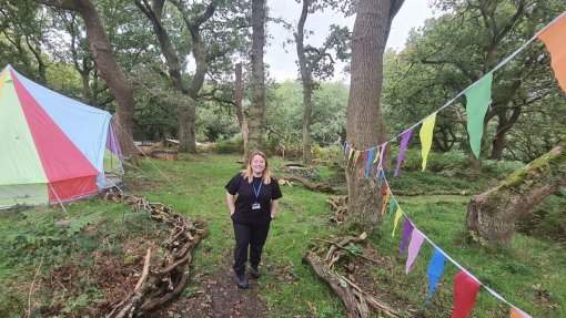 Menyw yn sefyll yn y goedwig gyda baneri'n hongian o'r coed a tipi lliwgar