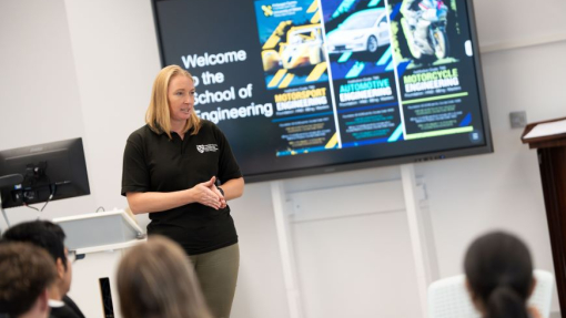 A lecturer wearing a branded top standing in front of a screen, watched by a classroom of sixth formers.