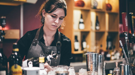 A girl making a cocktail surrounded by bottles at a bar.