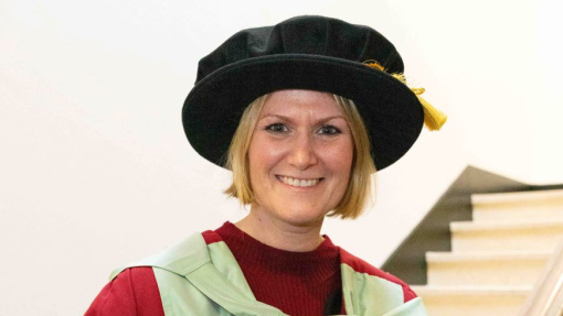 A smiling graduate dressed in her robes standing on a staircase.