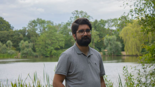 A happy smiling student standing close to a river with woodland behind.