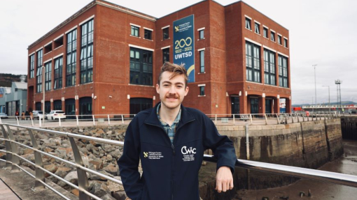 A male standing against a railing with a large red brick building in the background.