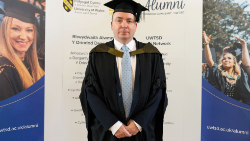 A happy graduate pictured in his robes against an alumni banner.