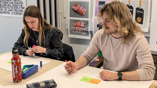 Two students with work displayed on a white table, sitting in a lecture room with artwork displayed on the walls.
