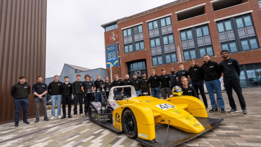 A large group of students standing proudly around a race car with the university building in the background.