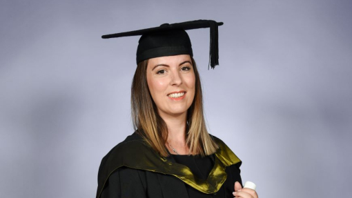 A happy smiling graduate dressed in her cap and gown holding a scroll tied with a red ribbon.