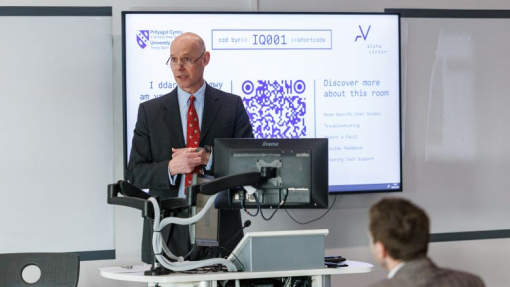 A man dressed in a suit standing in front of a white board, addressing students.