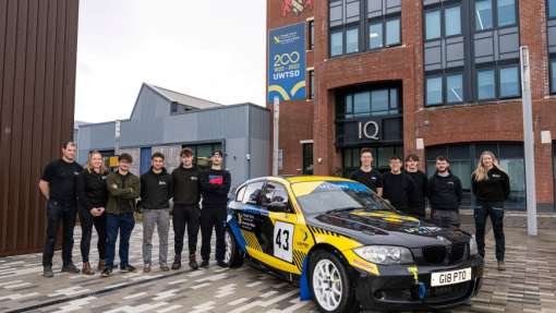 A group of motorsport students standing behind a yellow and black rally car in front of a university building.