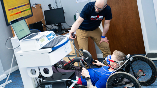 A man is on a hand-bike wearing a facemask for VO2 testing. Another man is crouching next to him holding up a device in eye view of the man on the hand-bike. A computer and computer screen is next to them monitoring the movement.