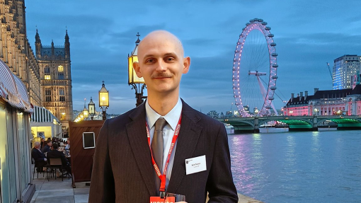 A man dressed in a suit holding a glass and wearing a visitor lanyard, standing outside the House of Commons in London.