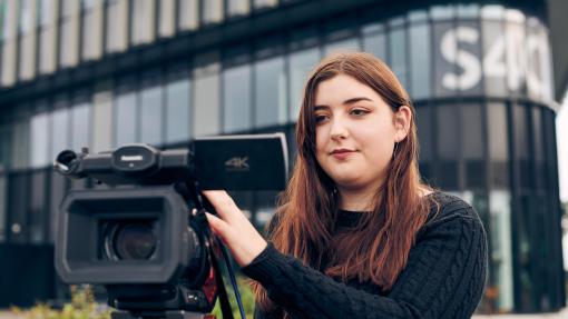 menyw yn sefyll y tu ôl i'r camera o flaen adeilad sy'n dweud S4C
