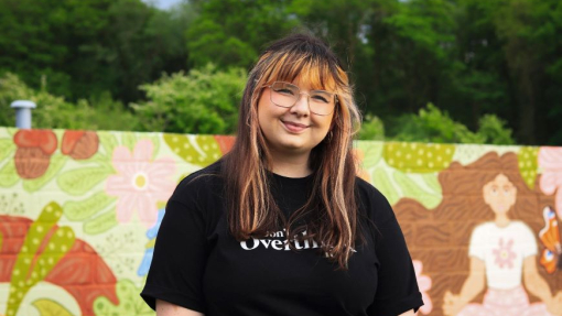 A smiling student dressed in a black t-shirt, standing in front of a brightly-coloured mural.