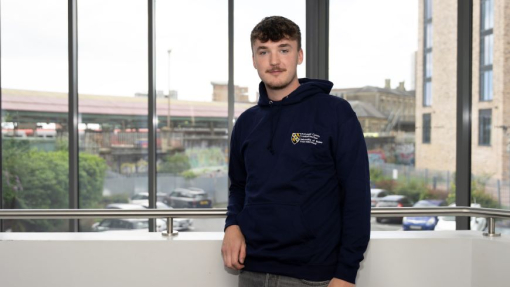 A male student standing in front of a large window, wearing a university branded hoodie.