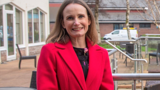 A pic of a smiling lady, dressed in a red coat, standing in front of university buildings.