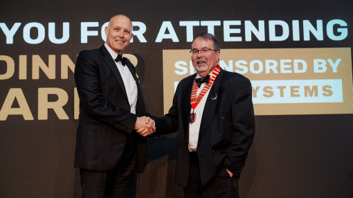 Two men dressed in black dinner suits with one holding an award, pictured against the backdrop of an events awards dinner.