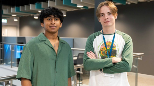 Two male students, one of whom has his arm folded, standing against a glass handrail.