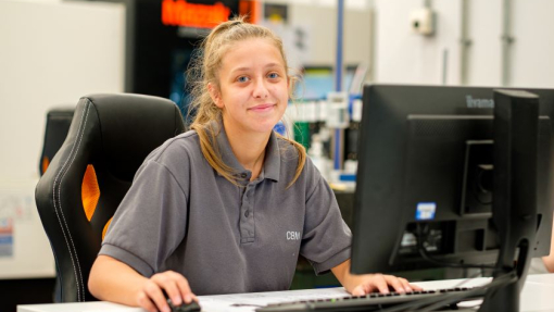 A woman dressed in a grey polo top sitting at a desk in a workshop environment.