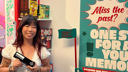 A smiling student in a white top holding an award and standing in front of her creative work.