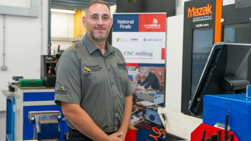 A man standing with his arms folded, wearing a branded top, in a workshop environment.