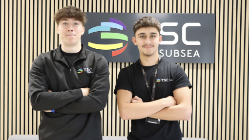 Two students dressed in black branded tops and trousers with their arms folded across their chests, standing in front of a grey sofa and a branded sign above.