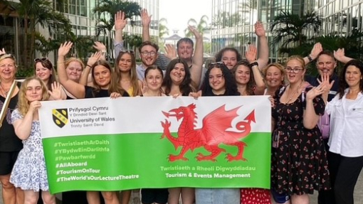A large group of students and staff holding a Welsh flag branded with the university's logo.