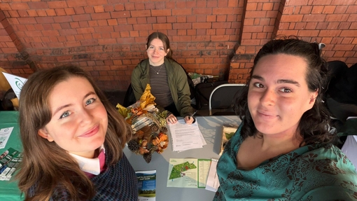 Three students, two of which are standing and one is seated, next to a table covered with leaflets and pens.