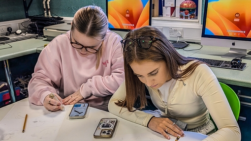 Two students sat a white desk working creatively in a lecture room.
