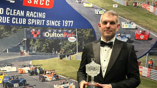 A man in a dinner suit holding a trophy in front of a racing branded banner.