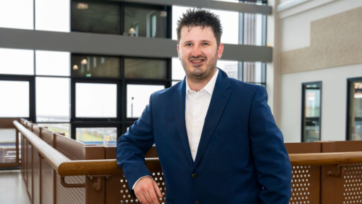 A graduate standing in front of a glass bannister on the top floor of a new building.